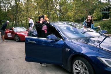 四名澳门威尼斯人网址的学生爬进了两辆特斯拉汽车的驾驶座和乘客座, 前景是一辆蓝色轿车，背景是一辆运动型红色跑车.